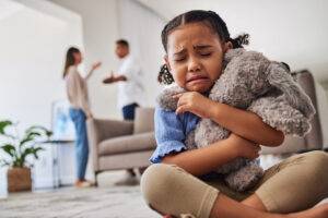 Trauriges, kleines Mädchen weint und umarmt Teddy zu Hause, Eltern streiten sich im Hintergrund