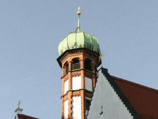 Ein historischer Turm in Augsburg ragt mit seiner grünen Kuppel und dem vergoldeten Ornament gegen den klaren Himmel, ein Symbol für die architektonische und religiöse Pracht und Geschichte der Stadt