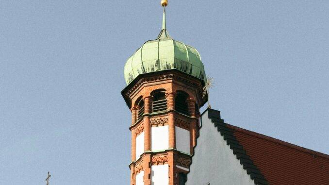 Ein historischer Turm in Augsburg ragt mit seiner grünen Kuppel und dem vergoldeten Ornament gegen den klaren Himmel, ein Symbol für die architektonische und religiöse Pracht und Geschichte der Stadt