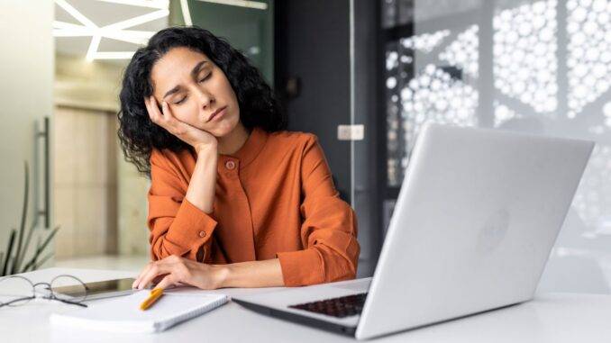 Frau in orangefarbener Bluse, die am Schreibtisch mit geschlossenen Augen und müdem Gesichtsausdruck auf ihre Hand gestützt ist, neben einem Laptop und Schreibutensilien.