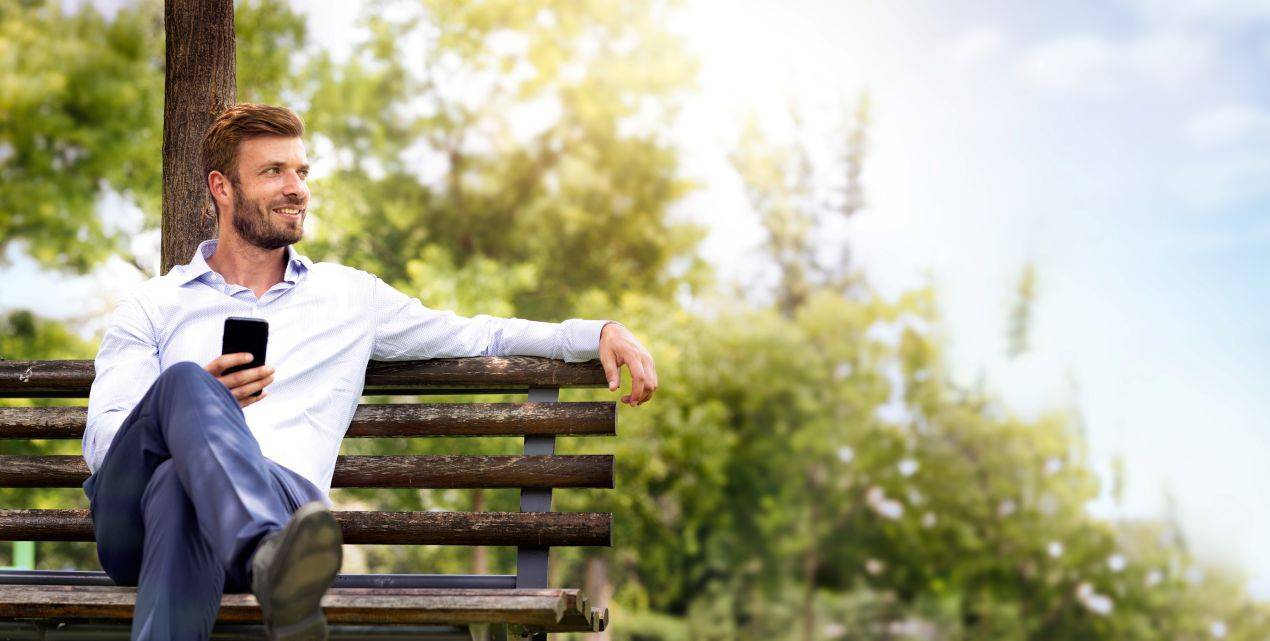 Mann in Business-Outfit sitzt auf einer modernen Parkbank und haelt ein Smartphone in der Hand bei sonnigem Wetter