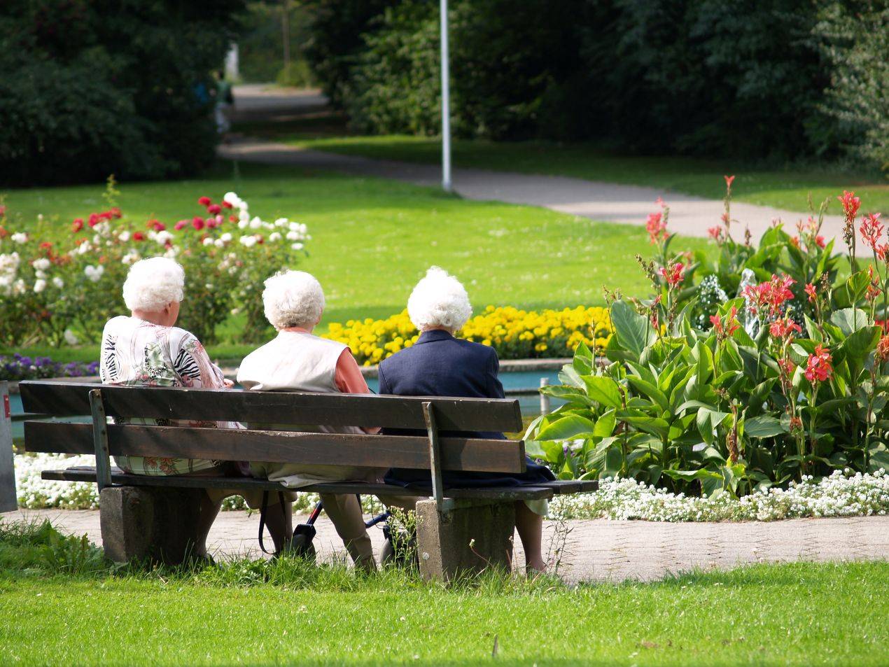 Drei aeltere Frauen sitzen auf einer Parkbank vor einem blumenreichen Beet in einem oeffentlichen Park